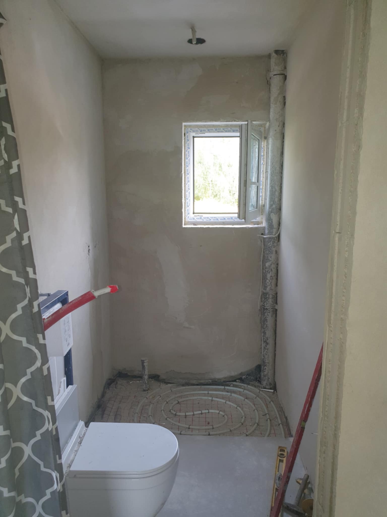 Łazienka w trakcie remontu: widoczna instalacja ogrzewania podłogowego z rurkami ułożonymi spiralnie, zamontowany stelaż podtynkowy WC, okno i surowe ściany.