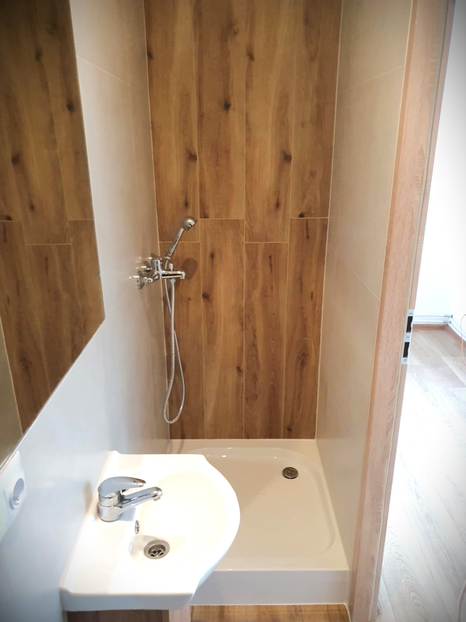 Nowoczesna kabina prysznicowa z umywalką, wykończona płytkami imitującymi drewno. Chromowana armatura i biała ceramika tworzą spójną całość.