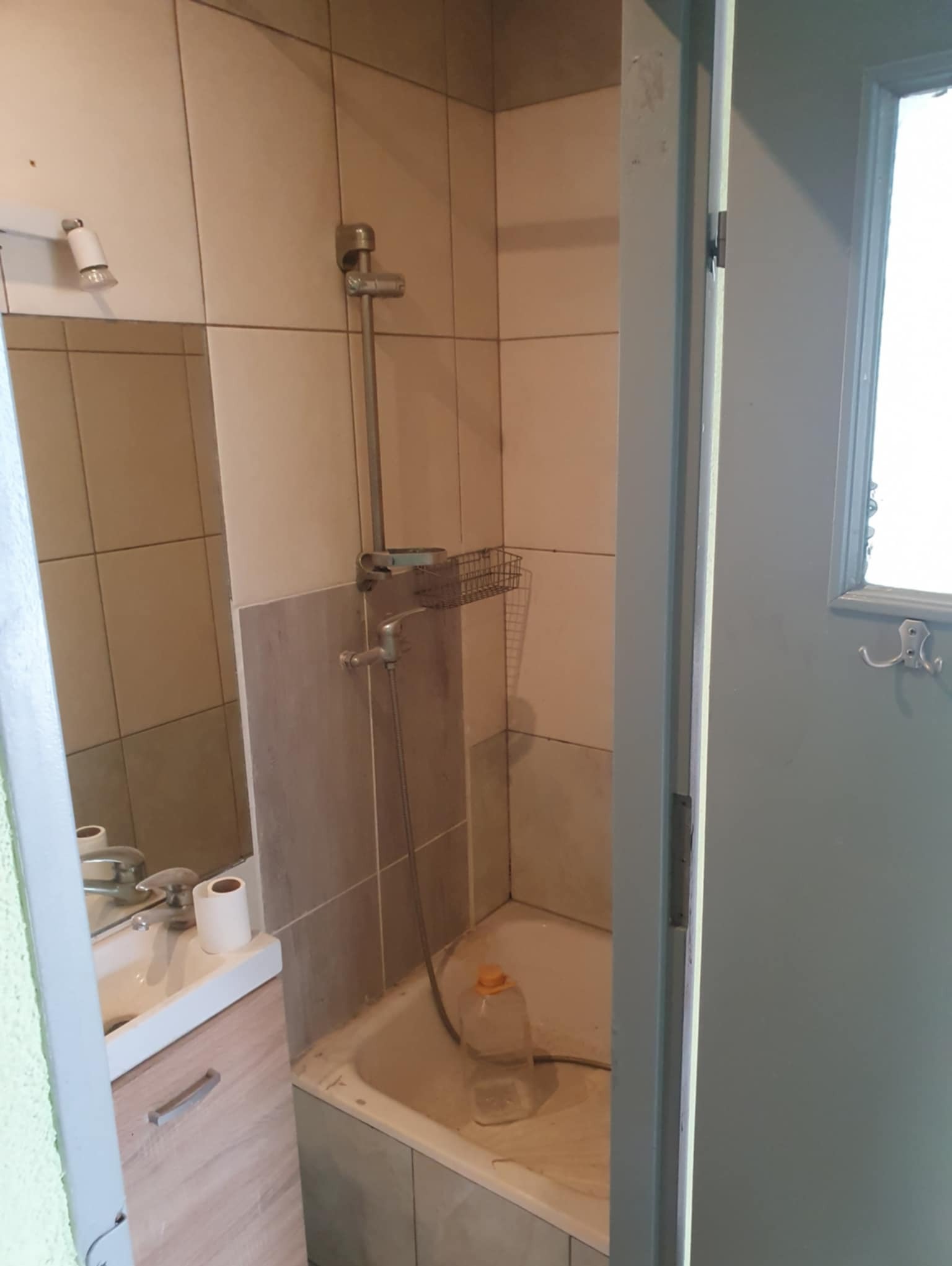 Umywalka z szafką, bateria i toaleta w łazience z beżowymi płytkami. Widoczny prysznic i drzwi. Wnętrze łazienki w trakcie remontu lub po nim.