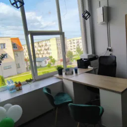 Jasne biuro z widokiem na osiedle przez duże okno. Na parapecie kwiaty, napoje i dekoracje. Biurko z krzesłami i sprzętem biurowym.