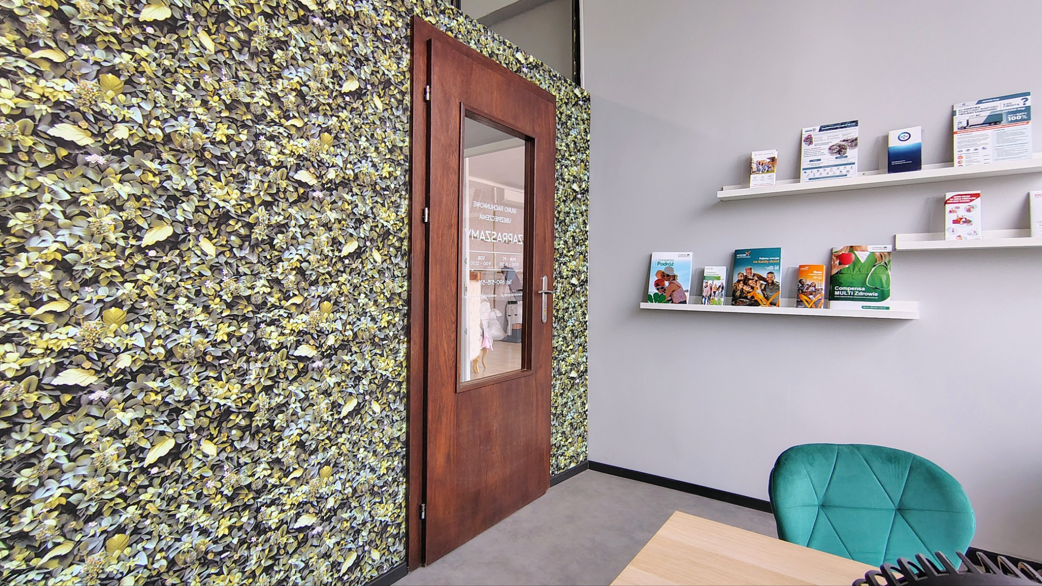 Wnętrze biura ubezpieczeniowego z zieloną ścianą, drewnianymi drzwiami z napisem 'Biuro Rachunkowe Ubezpieczenia' i półkami z broszurami informacyjnymi.