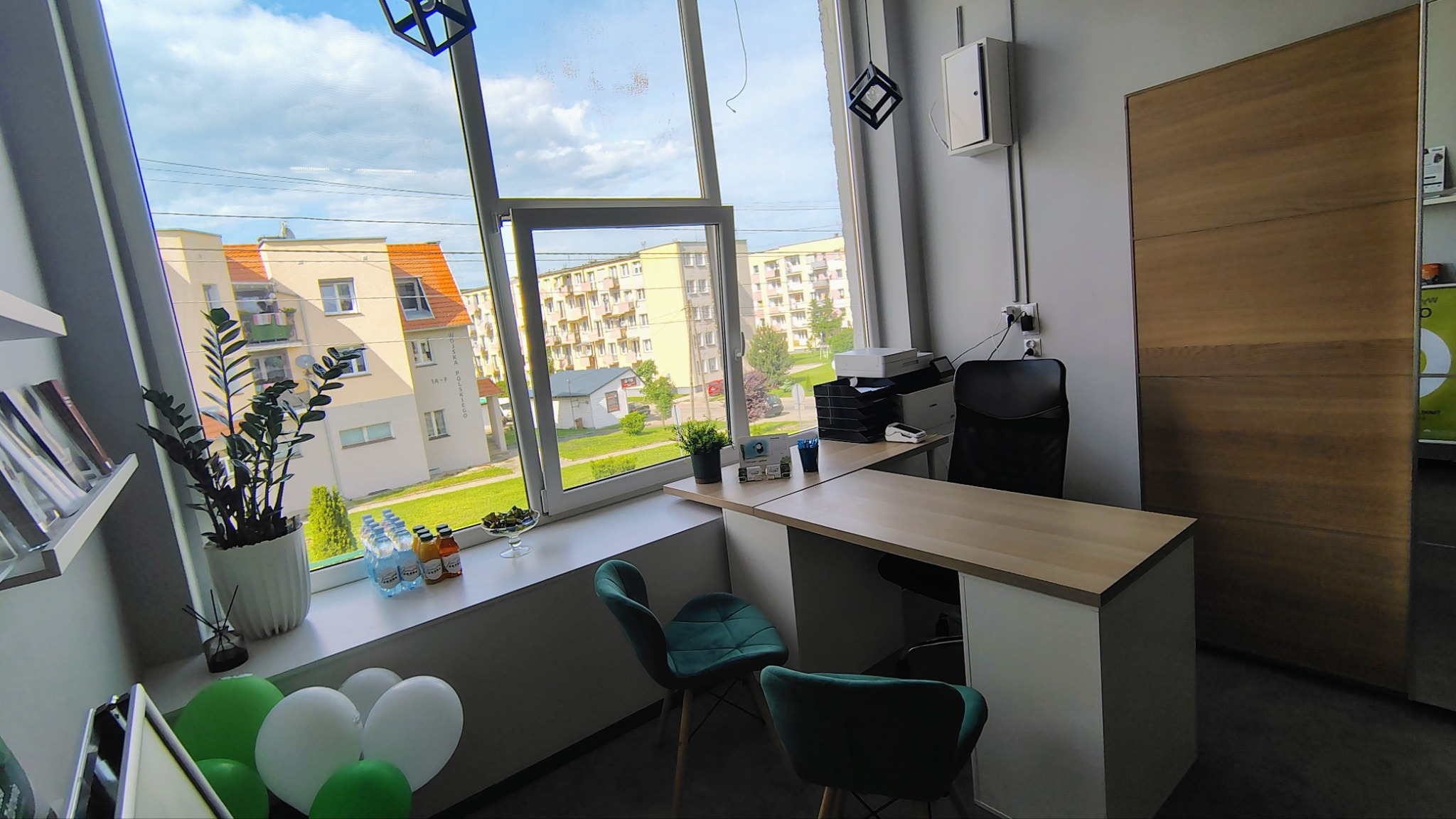 Jasne biuro z widokiem na osiedle przez duże okno. Na parapecie kwiaty, napoje i dekoracje. Biurko z krzesłami i sprzętem biurowym.