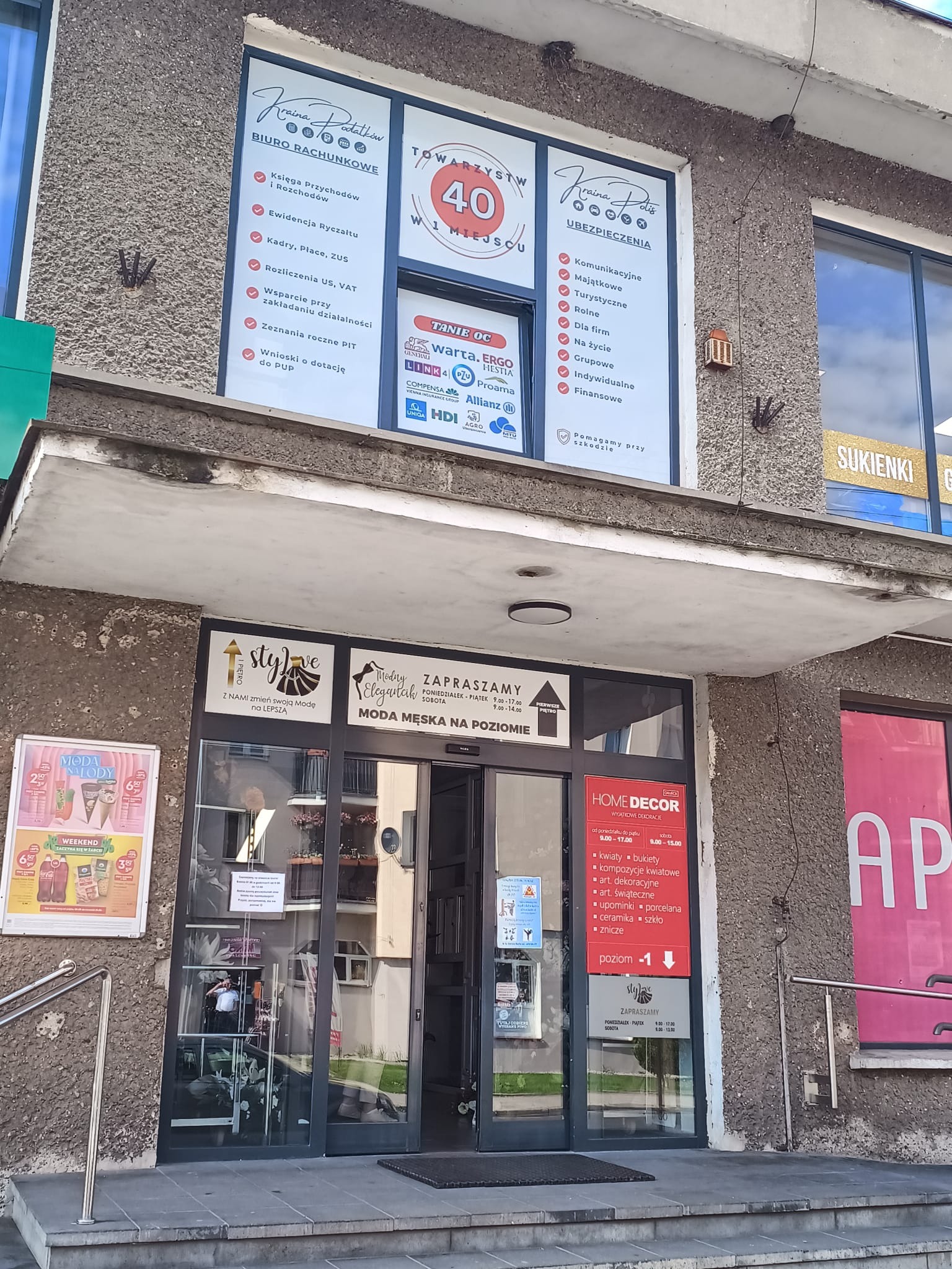 Fasada budynku z szyldami firm: biuro rachunkowe 'Kraina Podatków', ubezpieczenia, sklepy odzieżowe 'Stylove' i 'Modny Elegancik' oraz 'Home Decor'.