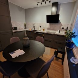 GLAZUROWSKY - Nowoczesna kuchnia w szarościach z okrągłym stołem i designerskim wazonem. Widoczne minimalistyczne szafki, oświetlenie punktowe i drewniana podłoga. Na stole leżą faktury.