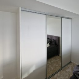 GLAZUROWSKY - Nowoczesna szafa wnękowa z przesuwnymi drzwiami, częściowo lustrzanymi, odbijająca fragment sypialni z łóżkiem. Minimalistyczny design, jasne kolory.