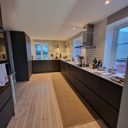 GLAZUROWSKY - Nowoczesna kuchnia z ciemnymi szafkami, jasnymi drewnianymi podłogami i dywanem. Na blacie akcesoria kuchenne i okno z widokiem na zewnątrz.