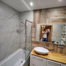 GLAZUROWSKY - Nowoczesna łazienka po remoncie: biała wanna z parawanem, umywalka nablatowa na szafce z drewnianym blatem, marmurowe i drewnopodobne płytki na ścianach.