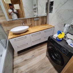 GLAZUROWSKY - Wykończona łazienka z drewnianym blatem, białą umywalką i szafkami. W tle lustro z odbiciem osoby robiącej zdjęcie. Na pralce leżą narzędzia i akcesoria.