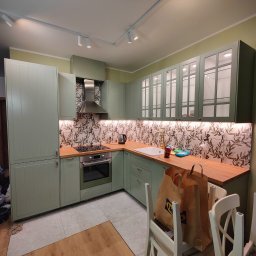 GLAZUROWSKY - Jasna kuchnia w odcieniach zieleni z drewnianym blatem i wzorzystymi płytkami. Widoczne oświetlenie podszafkowe i dwa krzesła z torbami IKEA.