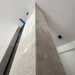 GLAZUROWSKY - Wykończony narożnik z szarych, marmurkowych płytek, z widocznymi niebieskimi zabezpieczeniami i czarnym profilem. Ujęcie z dołu, widoczny fragment sufitu z lampą.