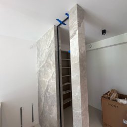 GLAZUROWSKY - Montaż kamiennej okładziny ściany działowej z ukrytymi drzwiami, zabezpieczonej niebieską taśmą. Widoczny fragment regału w prześwicie.