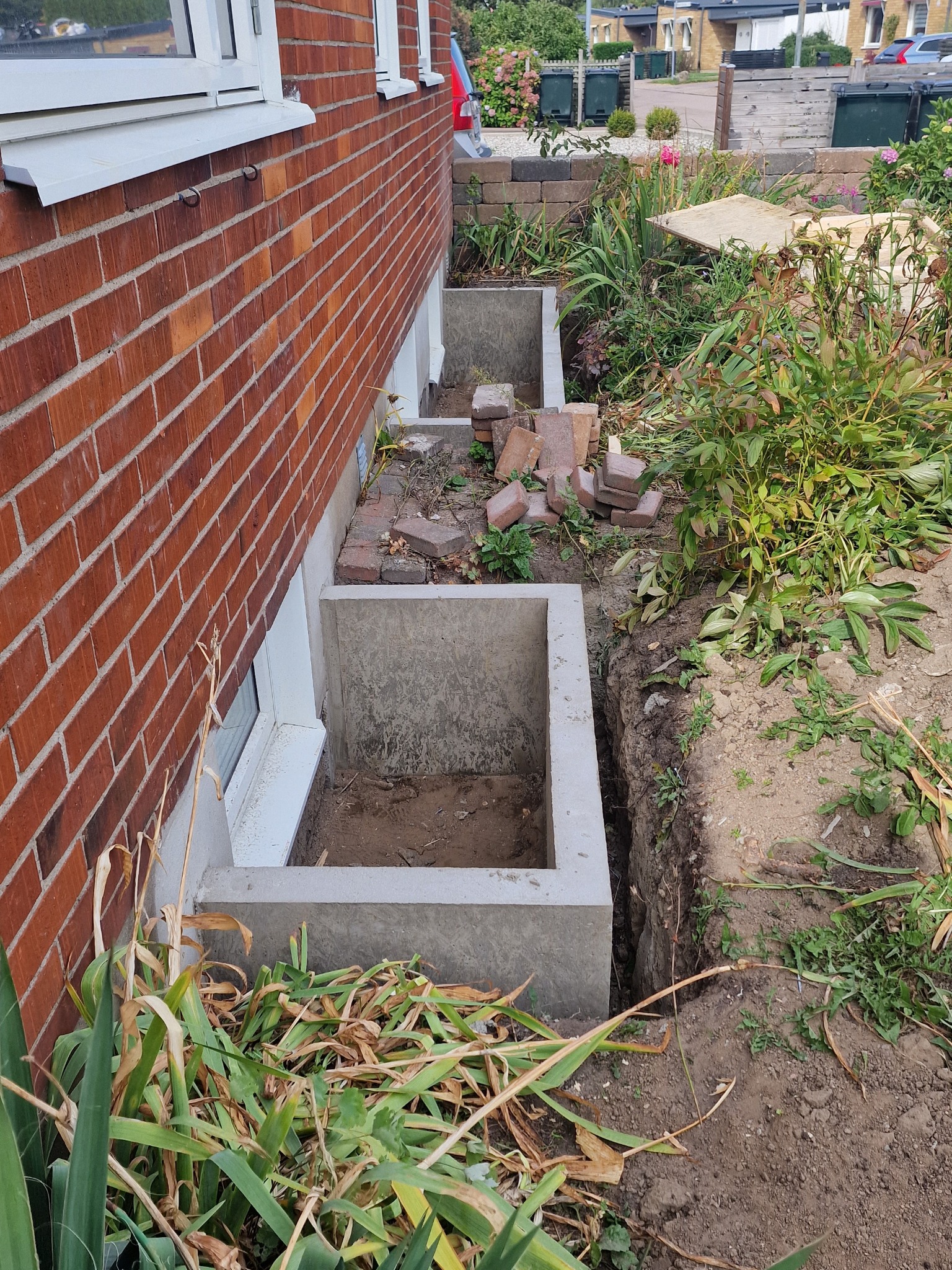 Betonowe obudowy okien piwnicznych przy ceglanym budynku, widoczne cegły do wykończenia i wykopana ziemia. Prace ziemne w toku, roślinność dookoła.