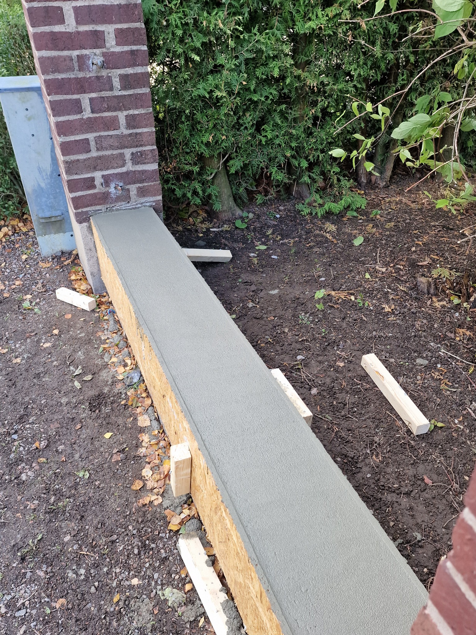 Świeżo wylana wylewka na drewnianym szalunku między dwoma ceglanymi słupkami w ogrodzie, z widocznymi drewnianymi podporami i zielenią w tle.