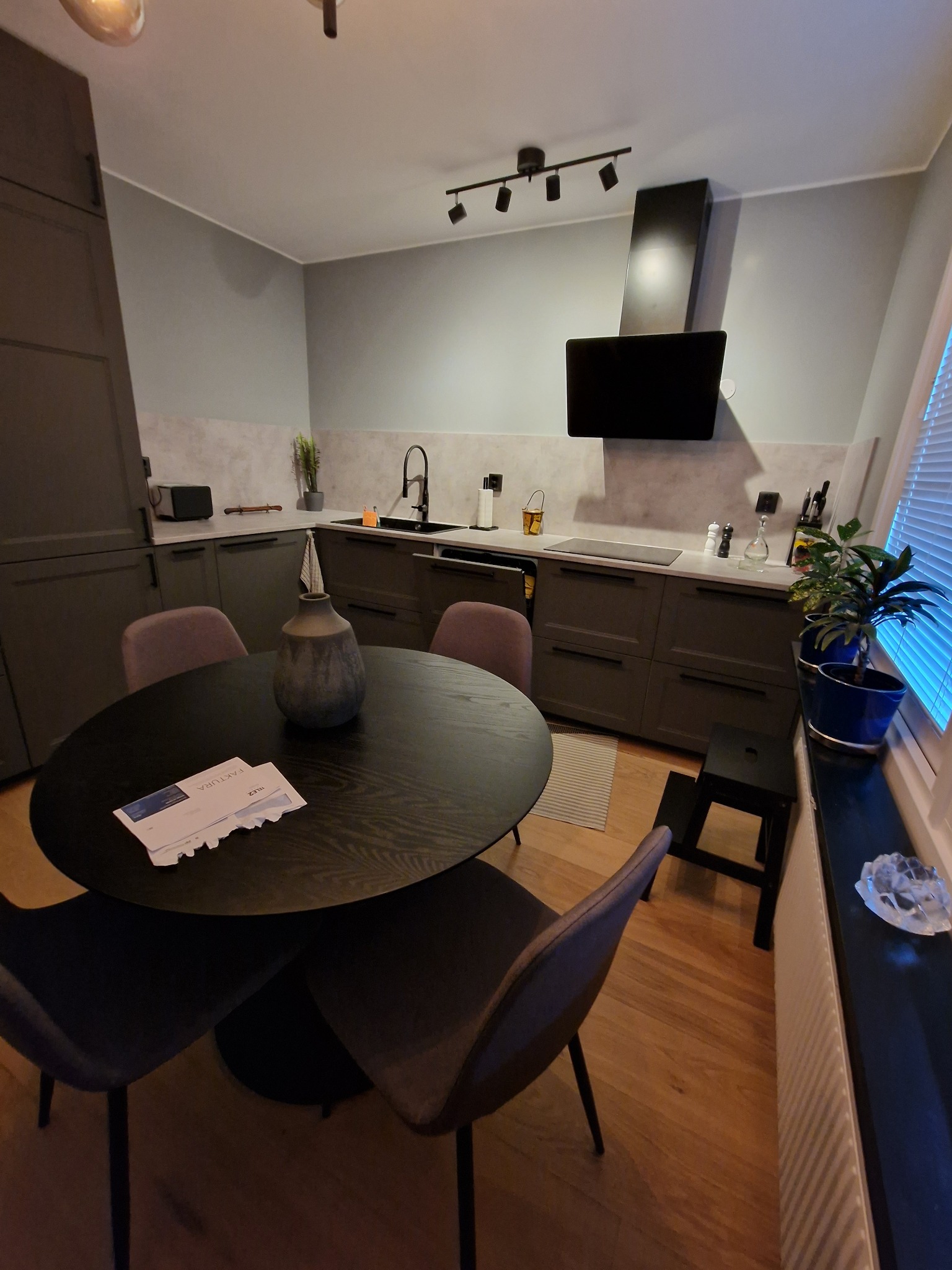 Nowoczesna kuchnia w szarościach z okrągłym stołem i designerskim wazonem. Widoczne minimalistyczne szafki, oświetlenie punktowe i drewniana podłoga. Na stole leżą faktury.