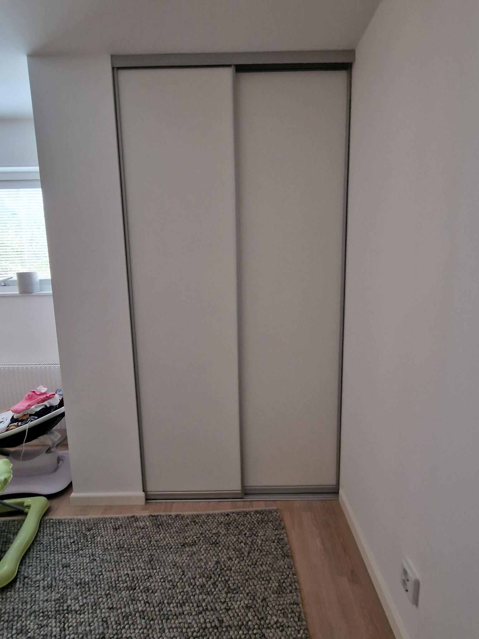 Biała szafa przesuwna z szarymi ramami, widoczna na tle jasnej ściany i fragmentu dywanu. W tle okno z żaluzjami.