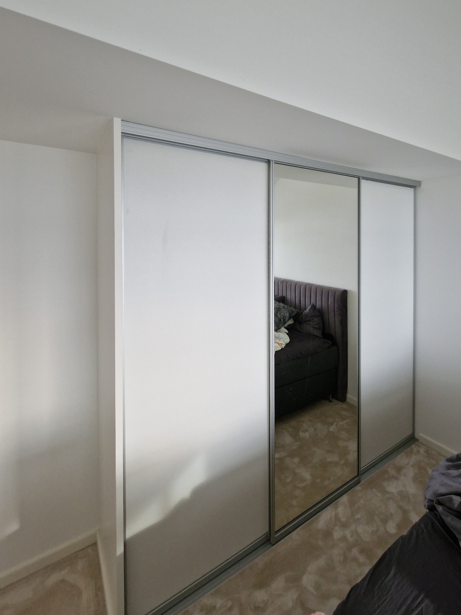 Nowoczesna szafa wnękowa z przesuwnymi drzwiami, częściowo lustrzanymi, odbijająca fragment sypialni z łóżkiem. Minimalistyczny design, jasne kolory.