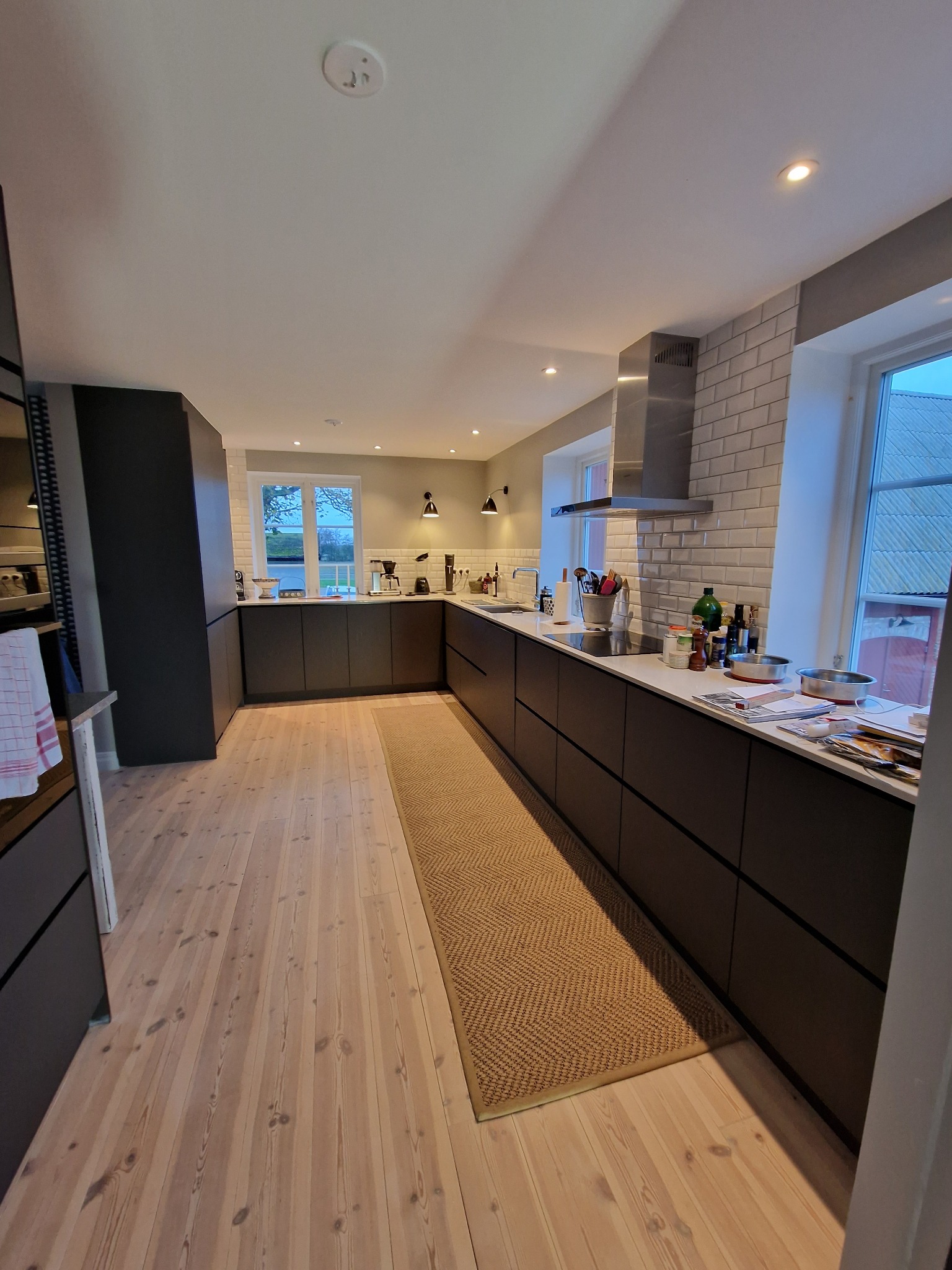 Nowoczesna kuchnia z ciemnymi szafkami, jasnymi drewnianymi podłogami i dywanem. Na blacie akcesoria kuchenne i okno z widokiem na zewnątrz.