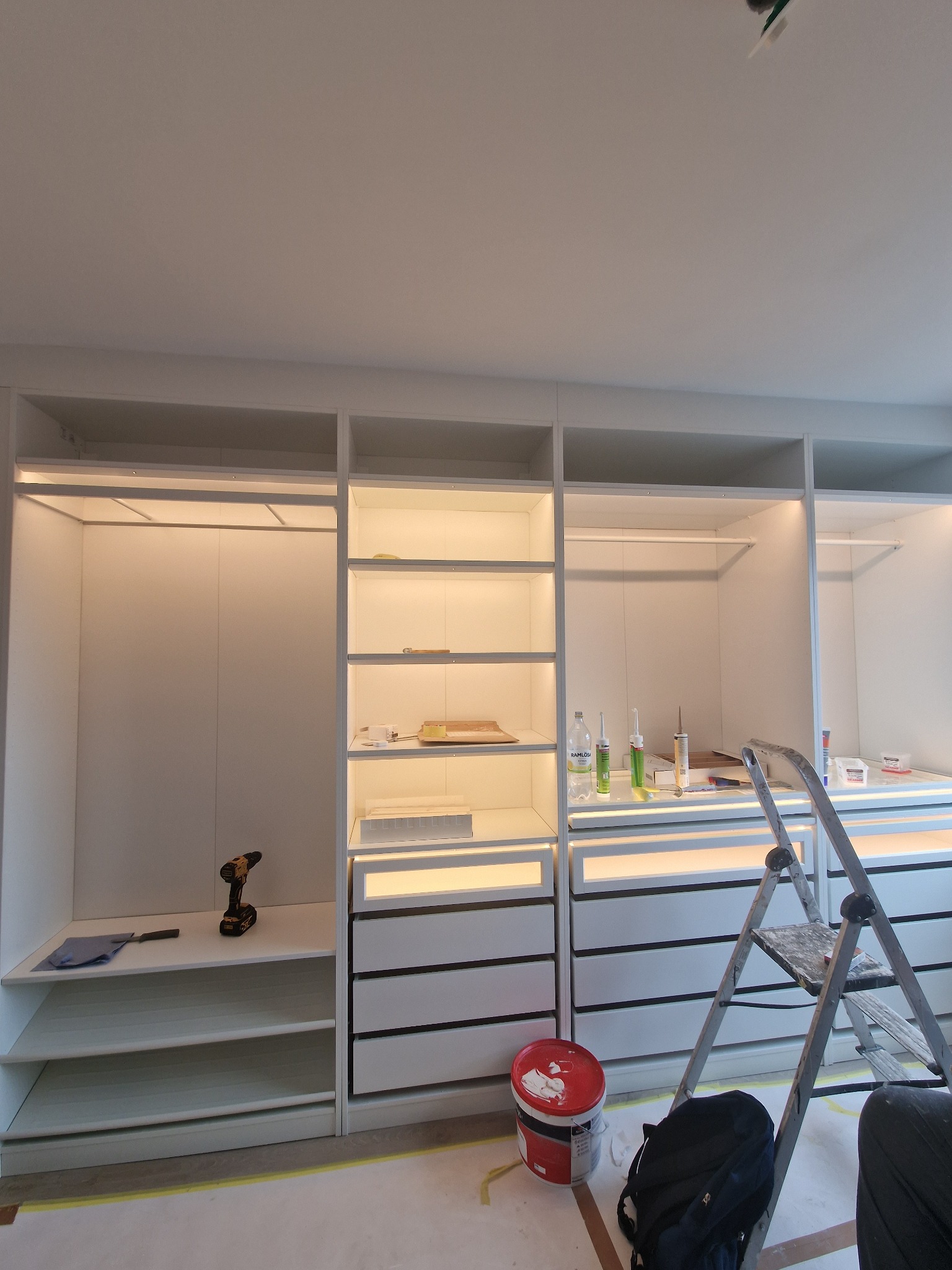 Biała, nowoczesna garderoba z oświetleniem LED, w trakcie montażu. Widoczna drabina, narzędzia i akcesoria monterskie. Ujęcie z dołu, podkreślające wysokość zabudowy.