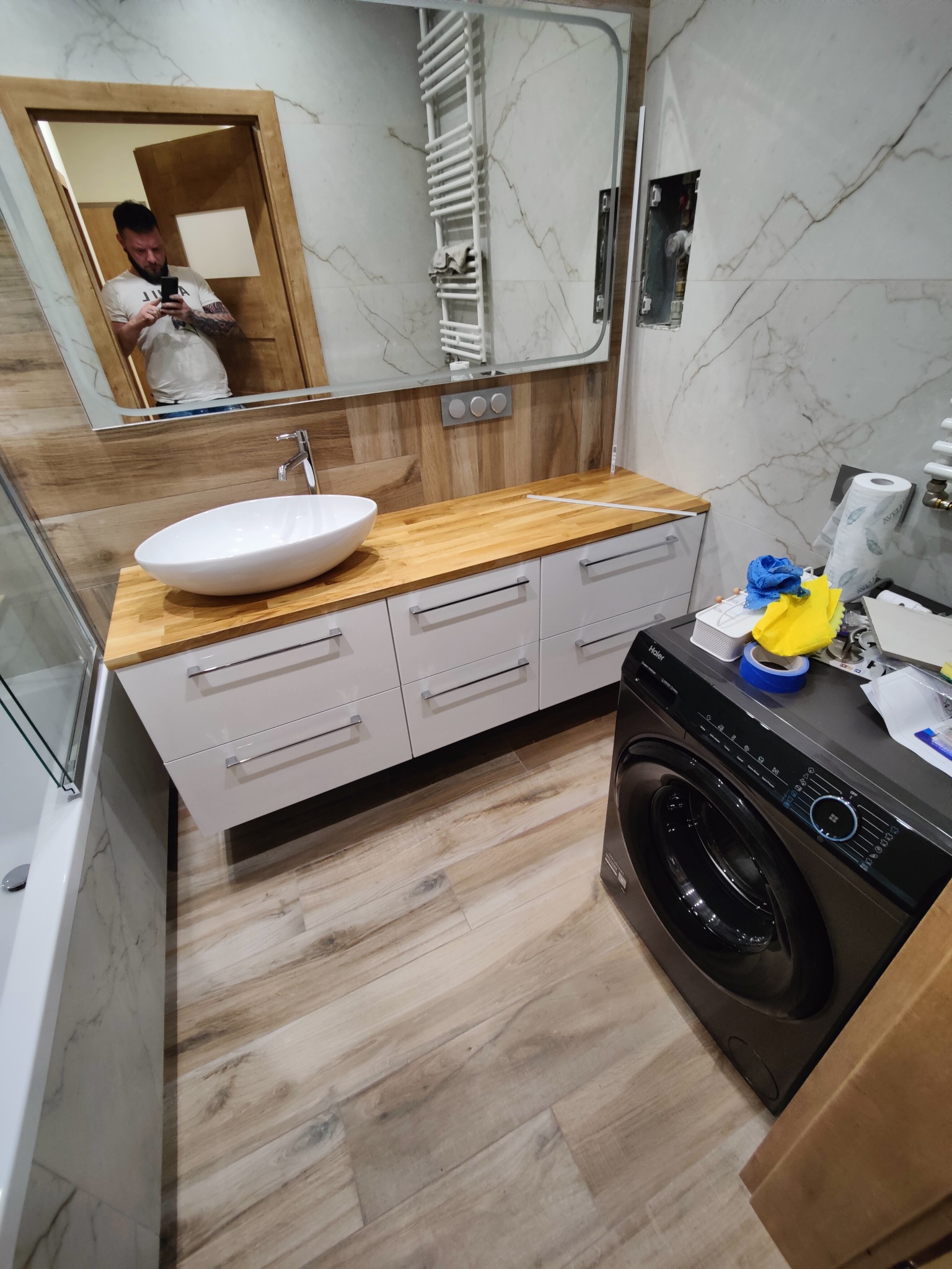 Wykończona łazienka z drewnianym blatem, białą umywalką i szafkami. W tle lustro z odbiciem osoby robiącej zdjęcie. Na pralce leżą narzędzia i akcesoria.