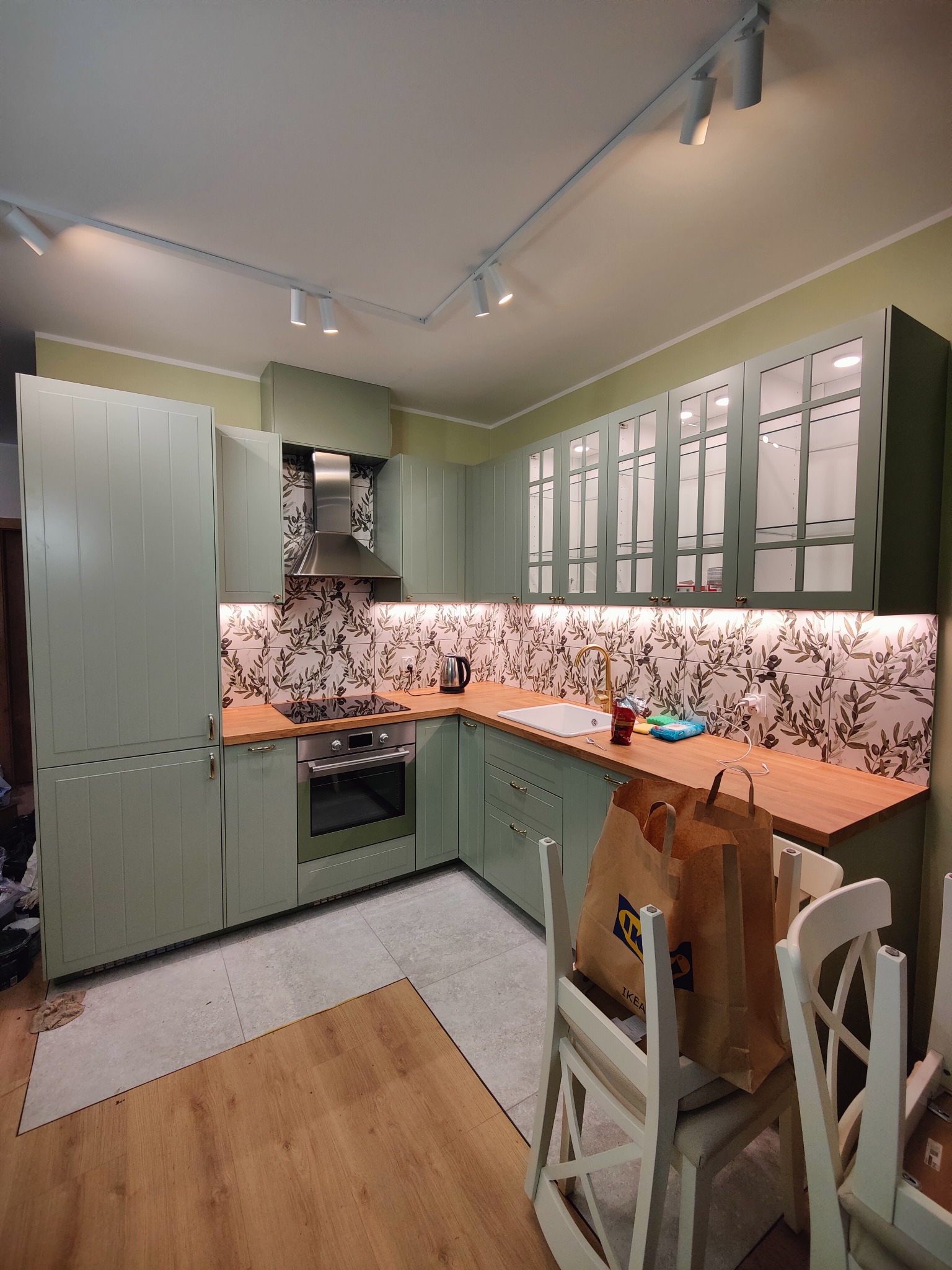 Jasna kuchnia w odcieniach zieleni z drewnianym blatem i wzorzystymi płytkami. Widoczne oświetlenie podszafkowe i dwa krzesła z torbami IKEA.