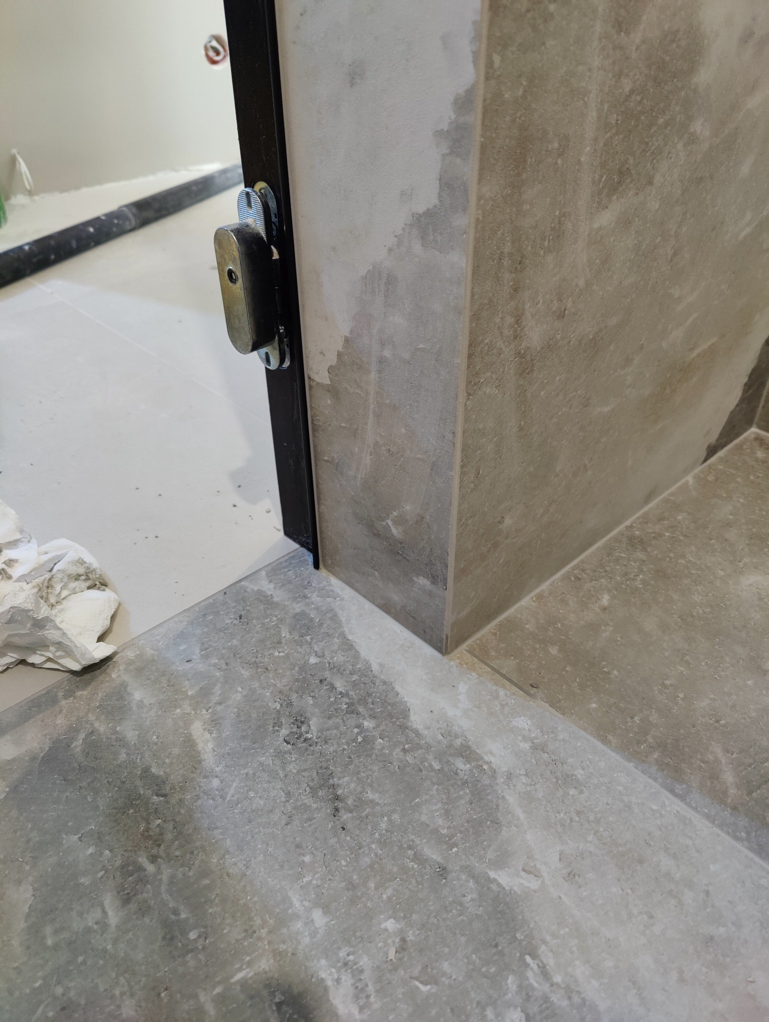 Fragment wykończonego wnętrza z nową, szarą, marmurkową podłogą z płytek i fragmentem ościeżnicy drzwiowej z zamkiem, ukazujący dbałość o detale.