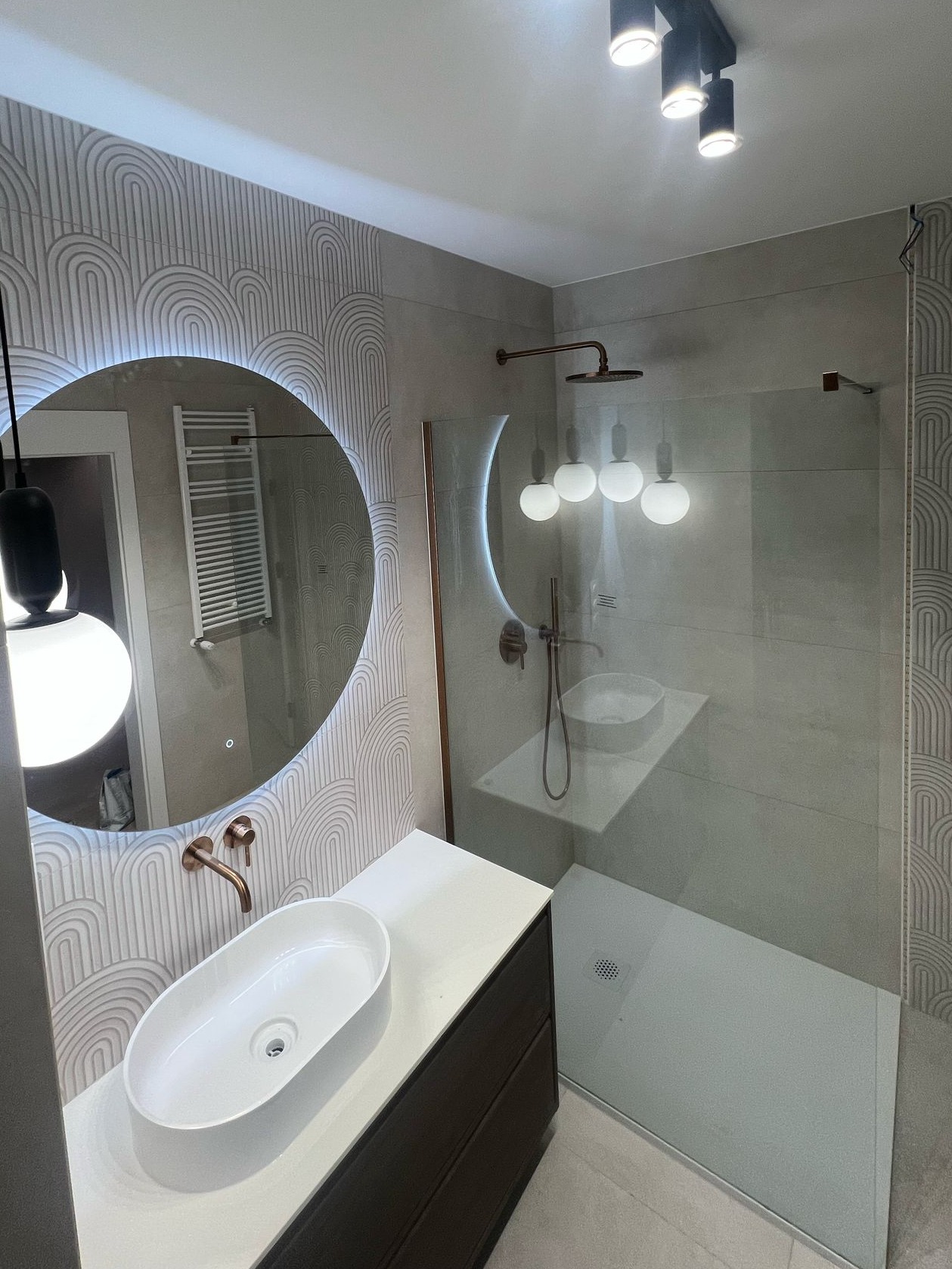 Nowoczesna łazienka z okrągłym lustrem LED, umywalką nablatową, kabiną prysznicową ze szklaną ścianką i armaturą w kolorze miedzi. Minimalistyczny design.