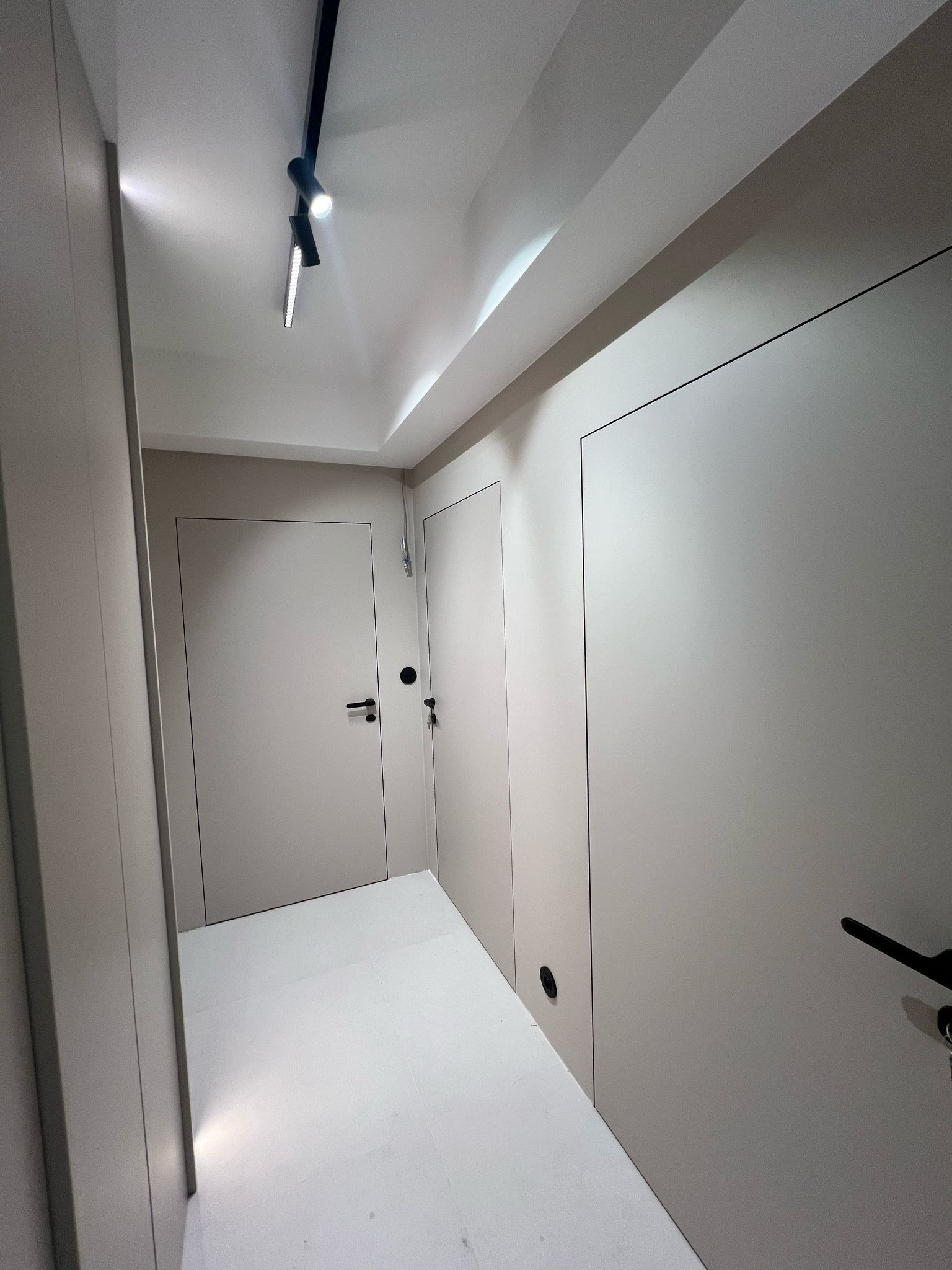 Minimalistyczny korytarz z szarymi, ukrytymi drzwiami i nowoczesnym oświetleniem punktowym. Czyste linie i jasna podłoga podkreślają prostotę wnętrza.