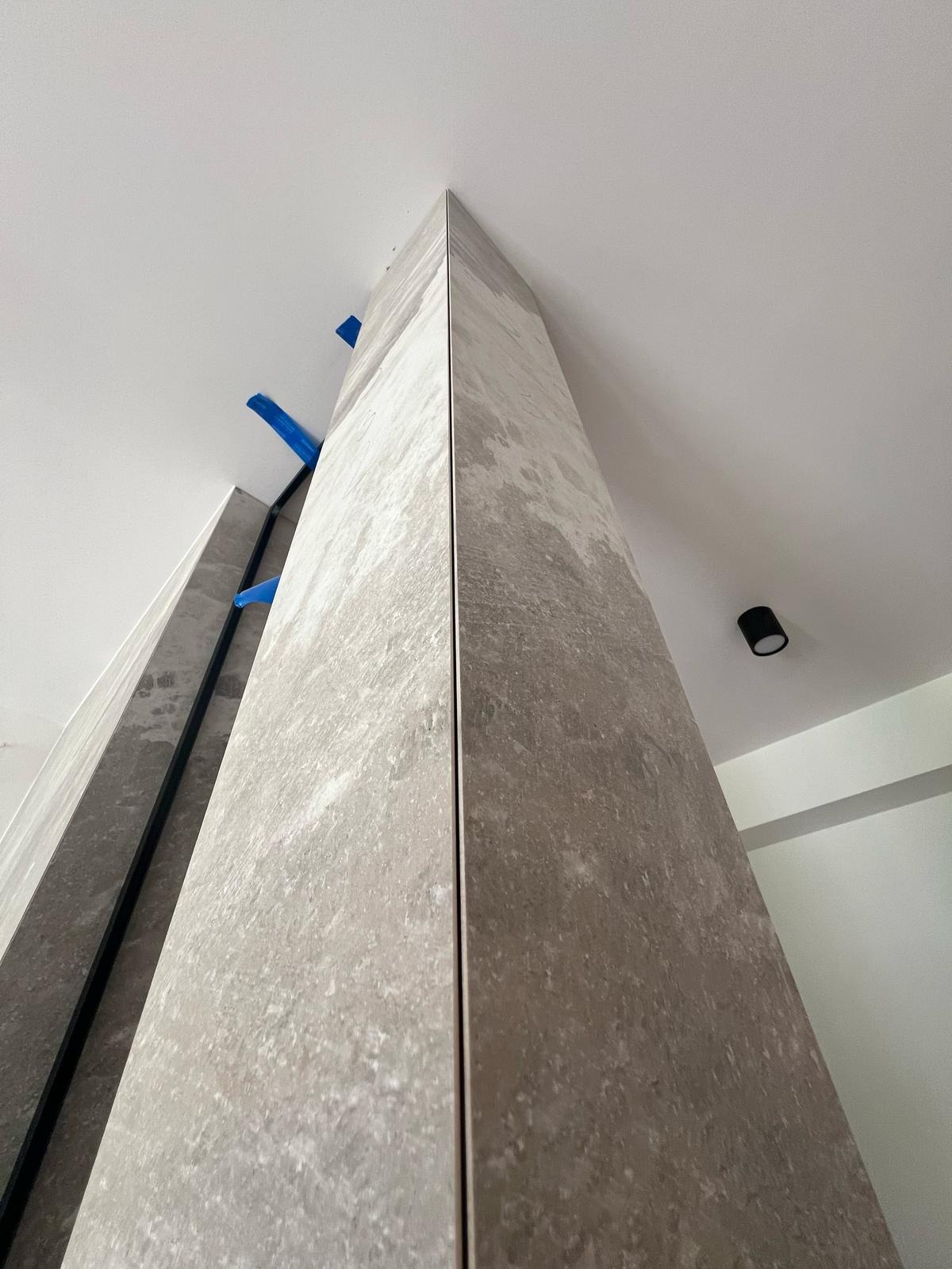 Wykończony narożnik z szarych, marmurkowych płytek, z widocznymi niebieskimi zabezpieczeniami i czarnym profilem. Ujęcie z dołu, widoczny fragment sufitu z lampą.