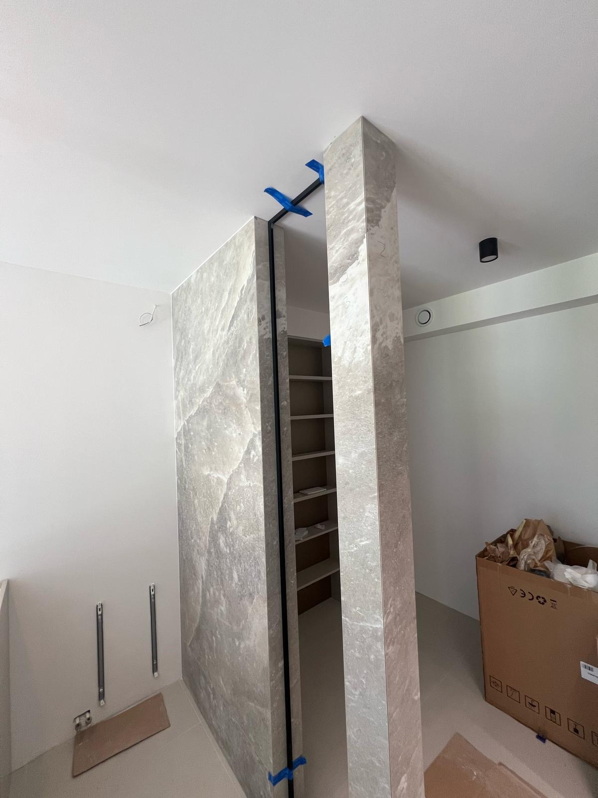Montaż kamiennej okładziny ściany działowej z ukrytymi drzwiami, zabezpieczonej niebieską taśmą. Widoczny fragment regału w prześwicie.