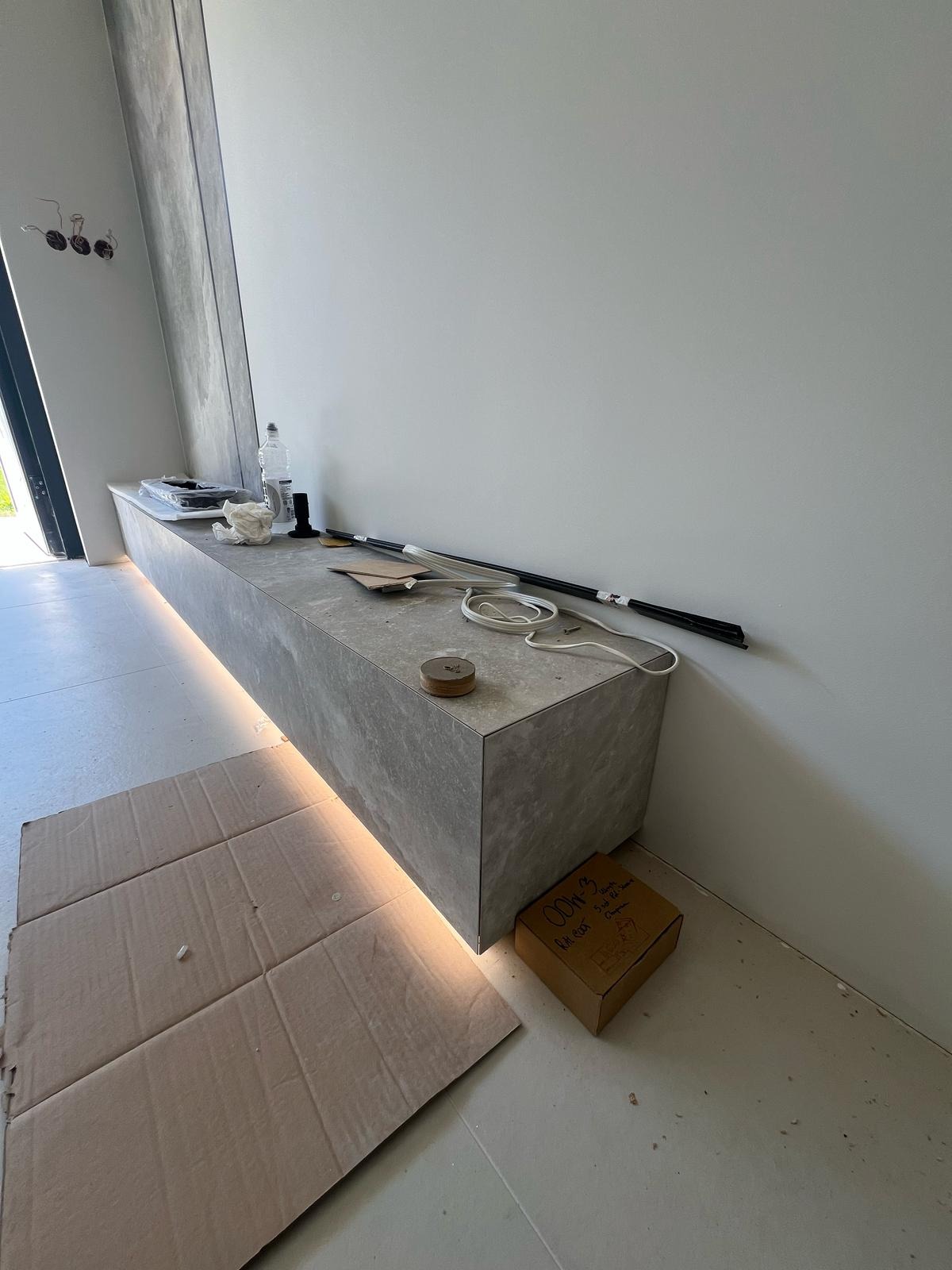 Szara, podświetlana listwą LED zabudowa wnęki z płytek. Na blacie narzędzia i akcesoria. Podłoga zabezpieczona kartonem, obok kartonowe pudełko.