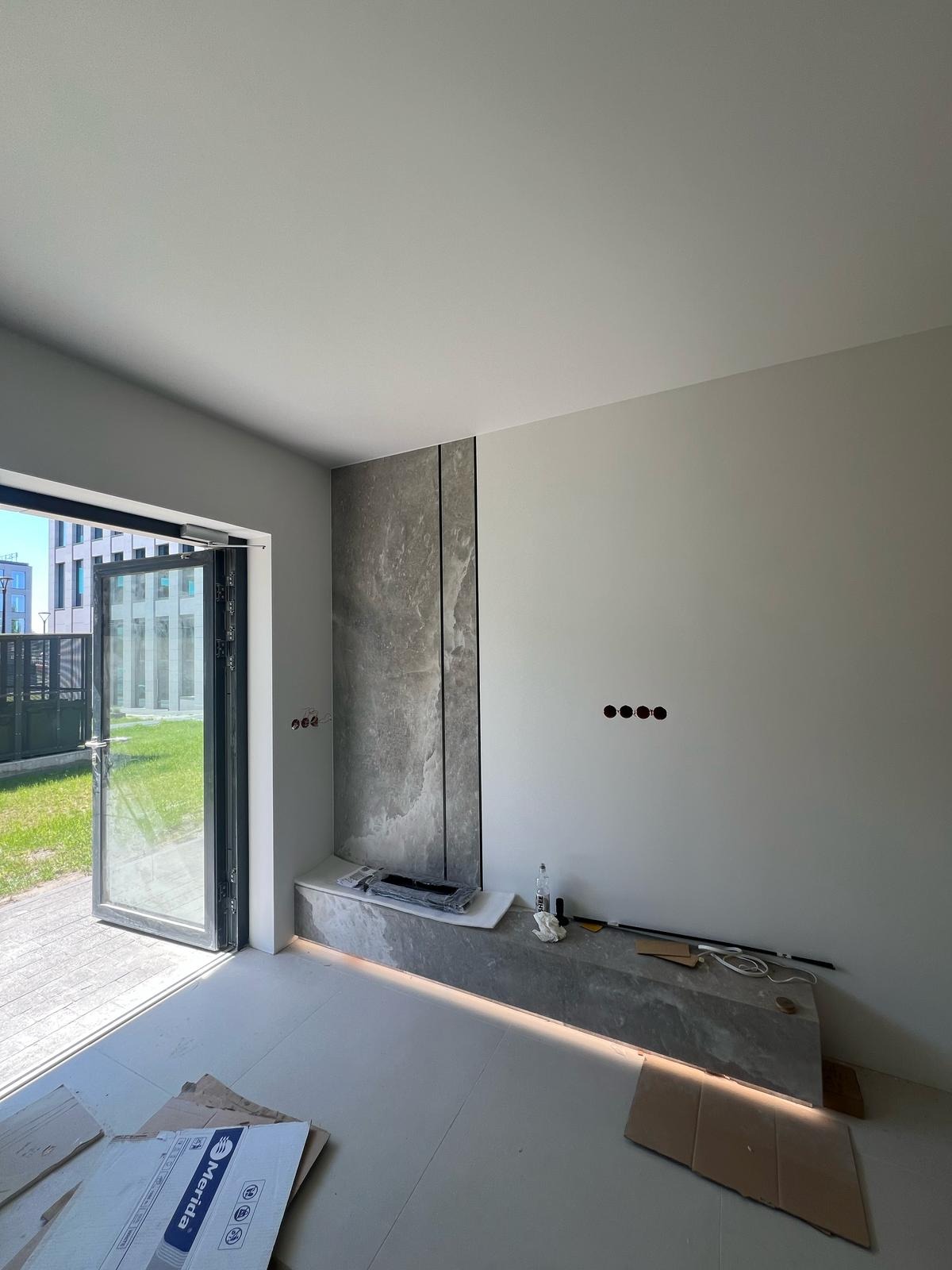 Wnętrze w trakcie wykańczania: szara ściana z marmurowym panelem, widoczne gniazdka i otwory na kable. Na podłodze płyty i kartony. Otwarty balkon z widokiem na zieleń.