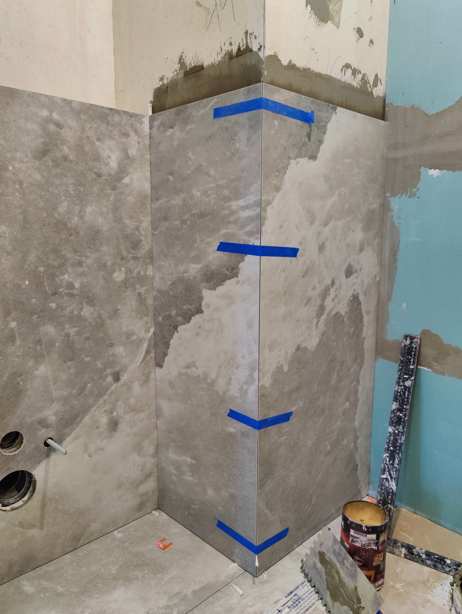 Wykończona ściana z szarych płytek imitujących marmur, zabezpieczonych niebieską taśmą w narożnikach. Widoczne narzędzia glazurnicze i fragment instalacji wodnej.
