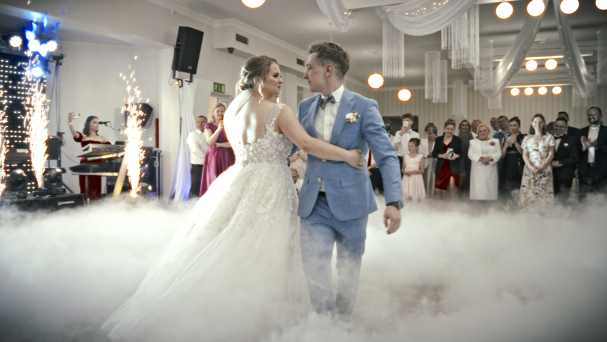 Para młoda w tańcu, otoczona dymem i fajerwerkami, w tle goście weselni. Panna młoda w sukni z koronkowymi detalami, pan młody w błękitnym garniturze.