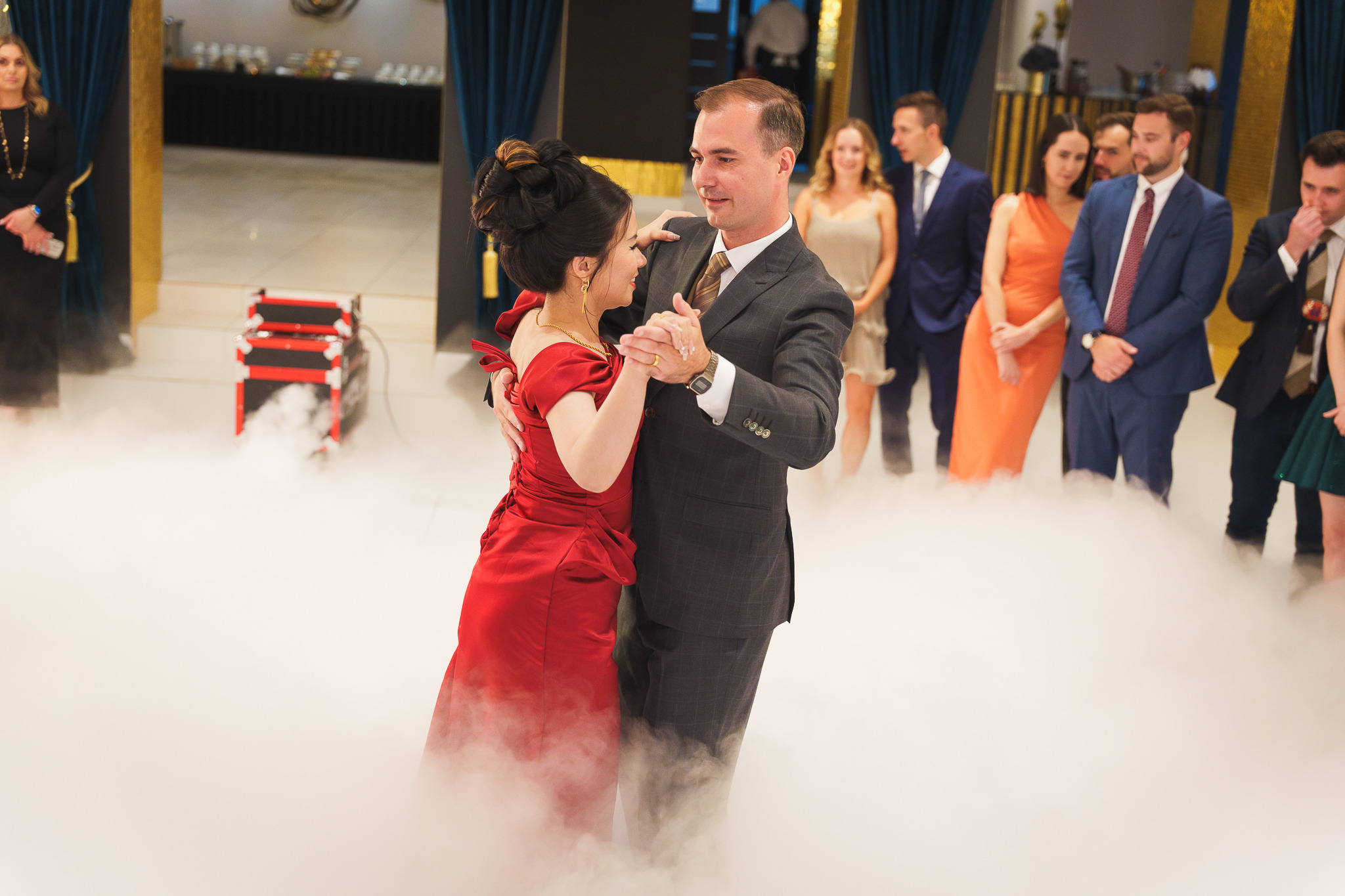 Para tańczy pierwszy taniec w chmurze dymu na weselu. Goście obserwują w tle. Kobieta w czerwonej sukni, mężczyzna w szarym garniturze. Dekoracje w kolorze granatowym i złotym.