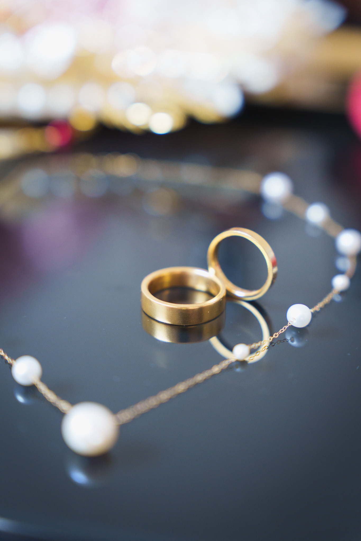 Dwa złote obrączki ślubne ustawione pionowo obok siebie na błyszczącej powierzchni z odbiciem, obok naszyjnik z pereł. Delikatne, rozproszone światło.