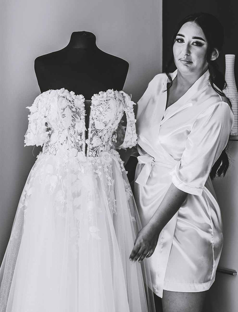 Panna młoda w szlafroku obok sukni ślubnej na manekinie. Delikatne zdobienia kwiatowe na sukni, satynowy szlafrok, stonowana kolorystyka. Przygotowania do ślubu.