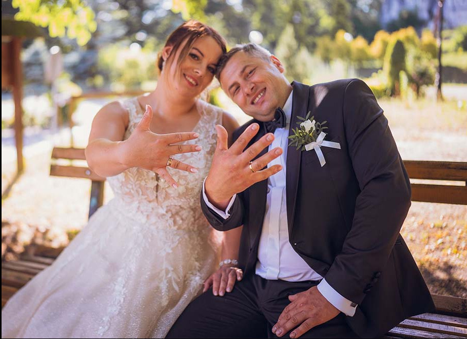 Uśmiechnięta para młoda siedzi na ławce, prezentując obrączki ślubne na tle zieleni parku w słoneczny dzień. Elegancki garnitur i suknia ślubna.