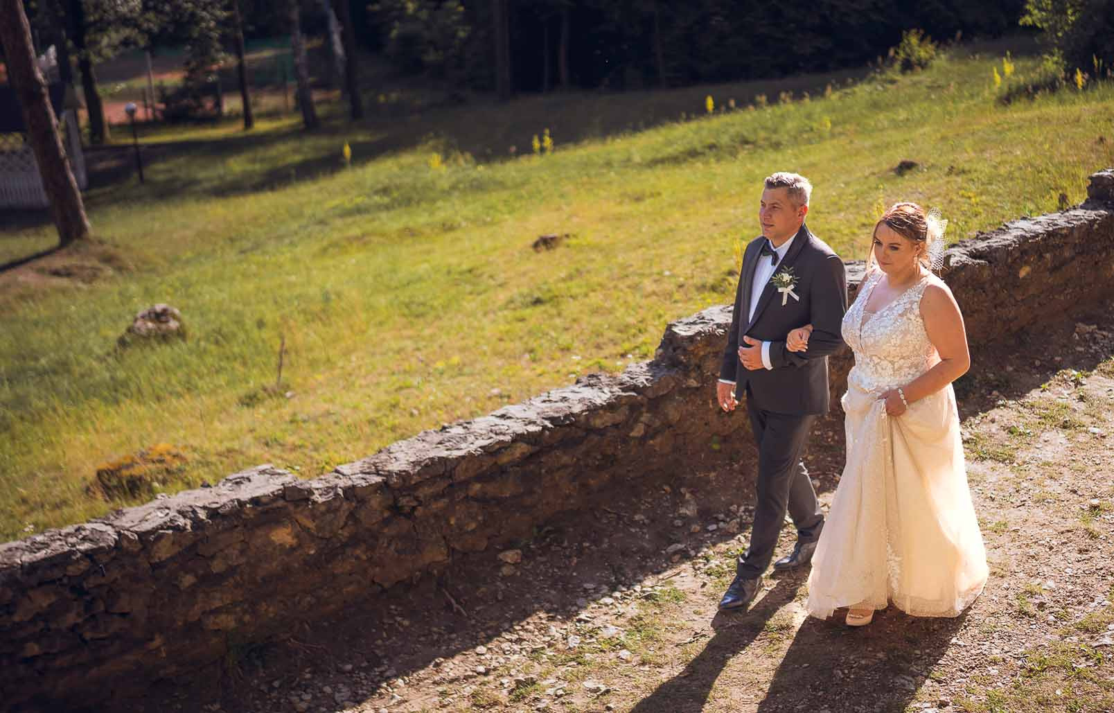 Para młoda spaceruje w słońcu po kamienistej ścieżce obok niskiego murku; on w szarym garniturze z butonierką, ona w koronkowej sukni ślubnej, trzymając ją lekko uniesioną.