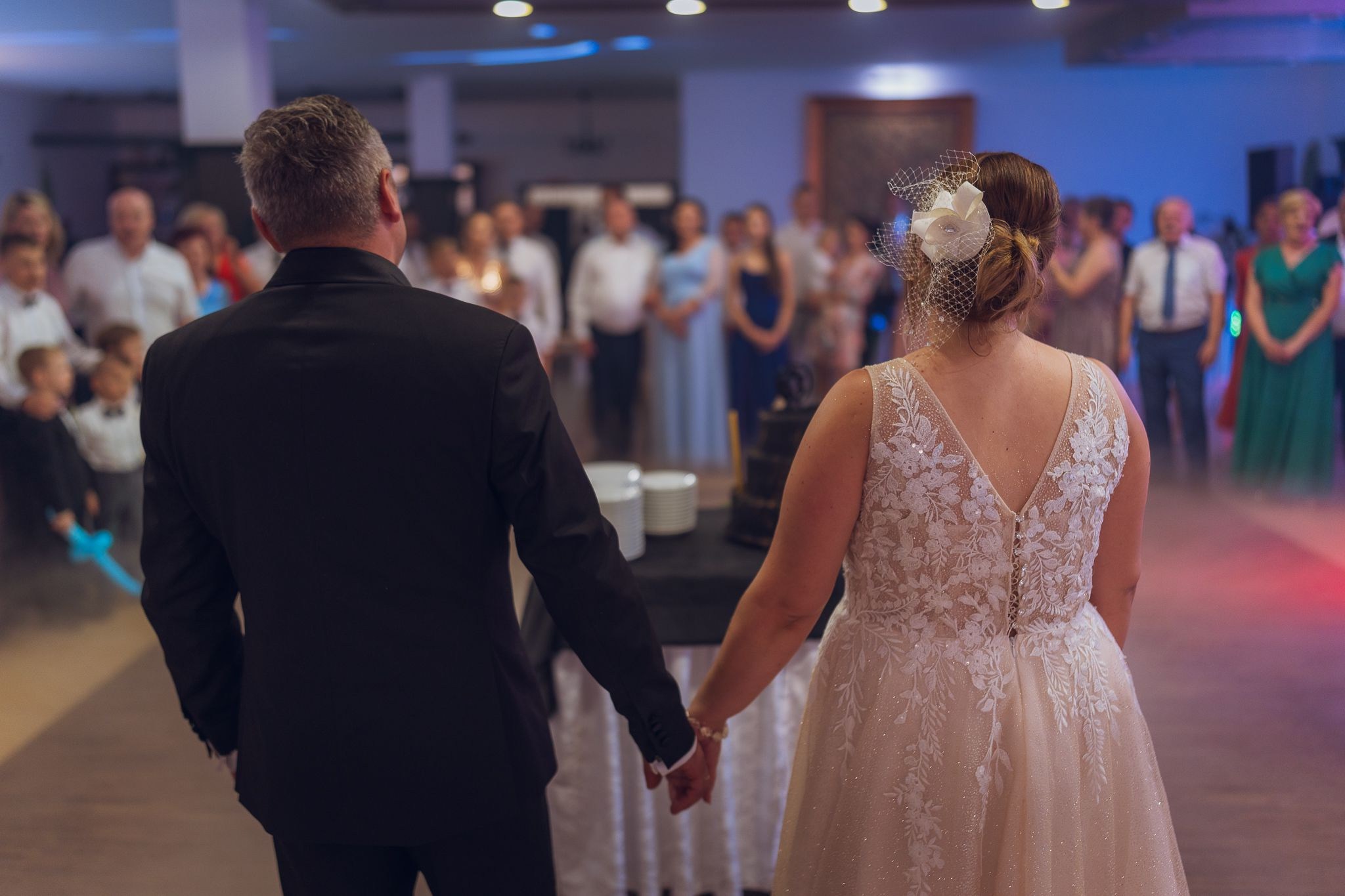Para młoda trzymająca się za ręce, tyłem do kamery, na sali weselnej z gośćmi w tle. Panna młoda w sukni z koronką, welon z kwiatem. Pan młody w czarnym garniturze.
