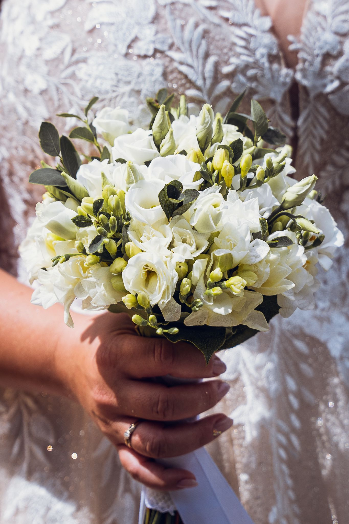 Panna młoda trzyma bukiet białych frezji i zielonych pąków na tle sukni ślubnej z koronką. Widoczny pierścionek zaręczynowy.