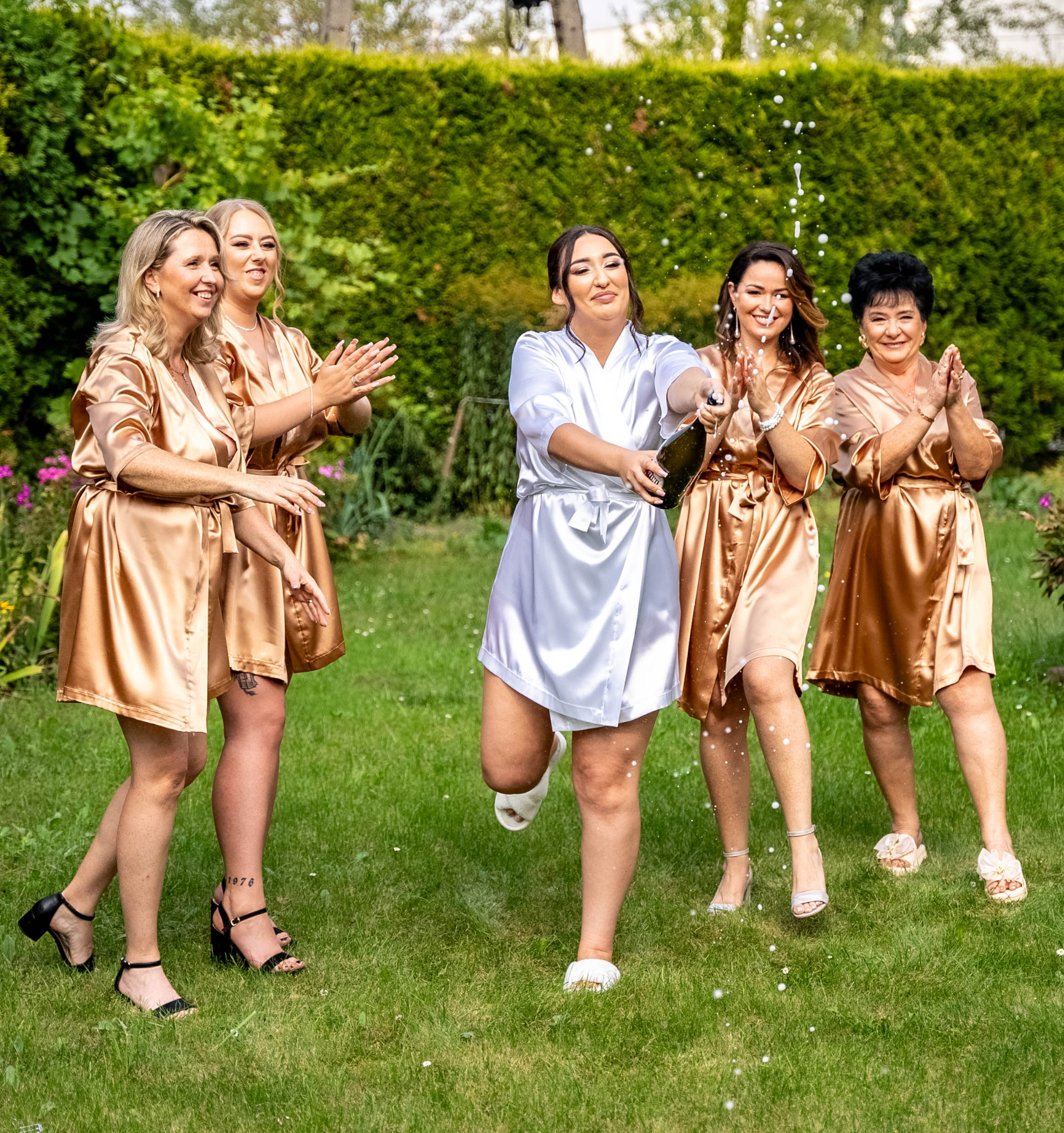 Panna młoda otwiera szampana w ogrodzie, otoczona przez druhny w szlafrokach. Radosna atmosfera, zielone tło z żywopłotu, dynamiczne ujęcie z fontanną tryskającego szampana.