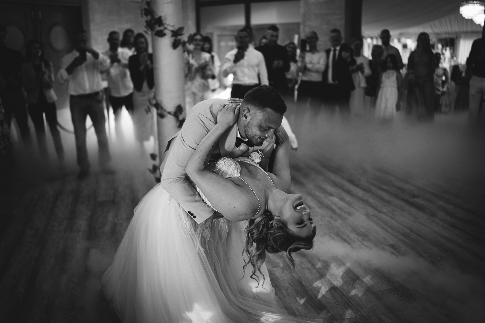 Czarno-białe zdjęcie pary młodej podczas pierwszego tańca w oparach dymu, z gośćmi weselnymi w tle. Panna młoda odchylona do tyłu, uśmiechnięta.