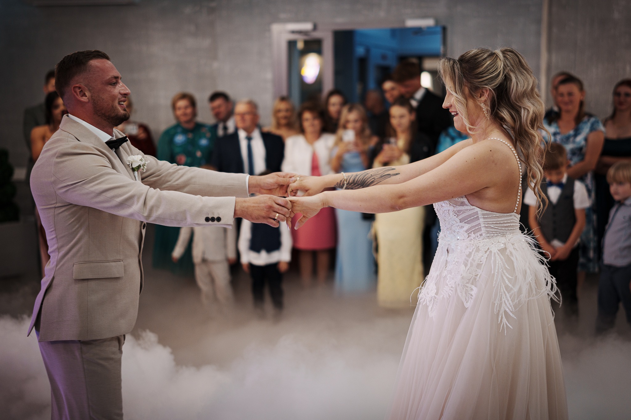 Para młoda w tańcu w chmurach dymu na tle gości weselnych. Panna młoda w sukni z piórami, pan młody w beżowym garniturze z muchą. Uśmiechnięci, wpatrzeni w siebie.