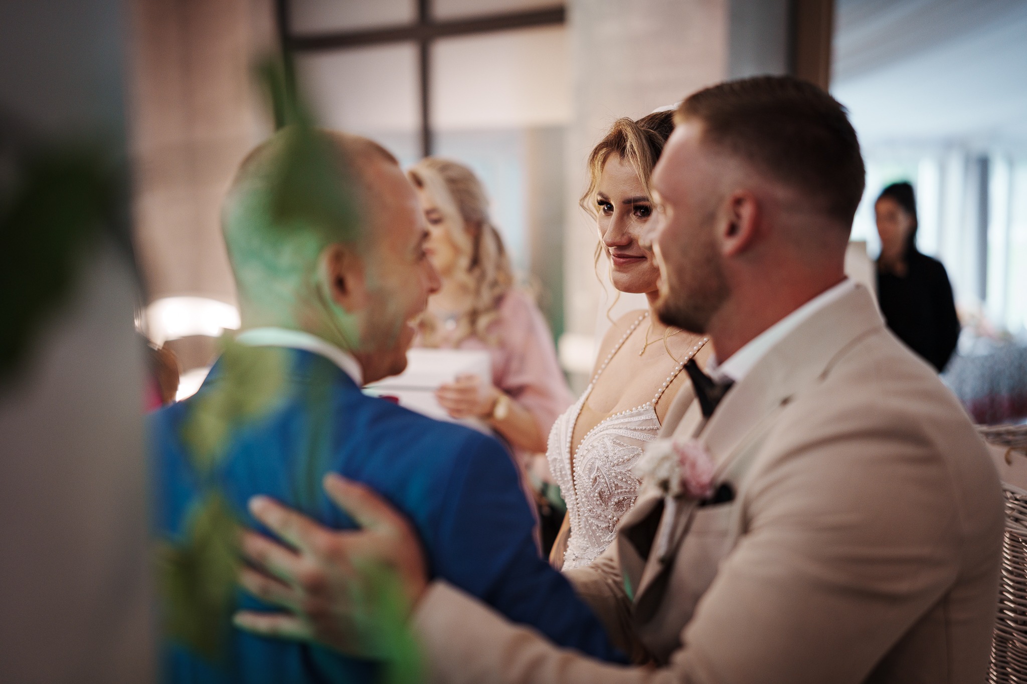 Uśmiechnięta panna młoda w koronkowej sukni spogląda na pana młodego w beżowym garniturze, w tle goście weselni. Delikatne, naturalne światło.