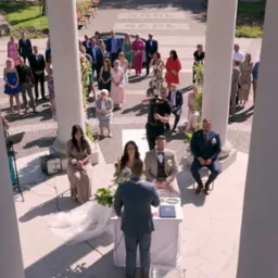 Uroczystość ślubna w plenerze, widok z góry przez kolumnadę, goście, para młoda, kamerzysta filmuje,  dekoracje kwiatowe.