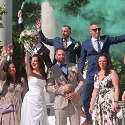 Pan Kamera - Uśmiechnięci goście weselni świętują z parą młodą na tle kolumn z kwiatami i kolorowych dymów.