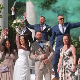 Uśmiechnięci goście weselni świętują z parą młodą na tle kolumn z kwiatami i kolorowych dymów.