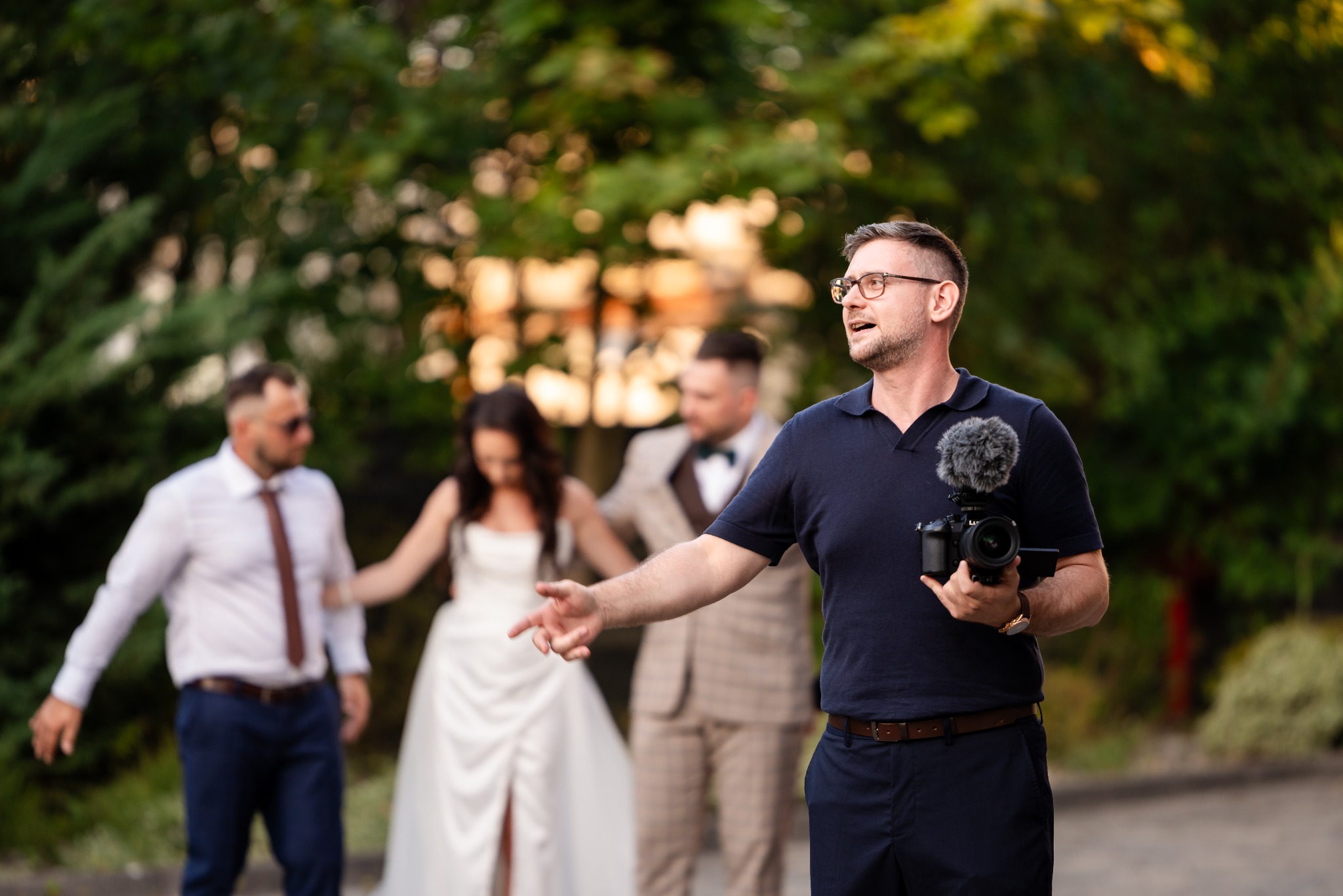 Kamerzysta z aparatem i mikrofonem reżyseruje parę młodą z gośćmi w tle, podczas plenerowej sesji ślubnej w zielonym otoczeniu.