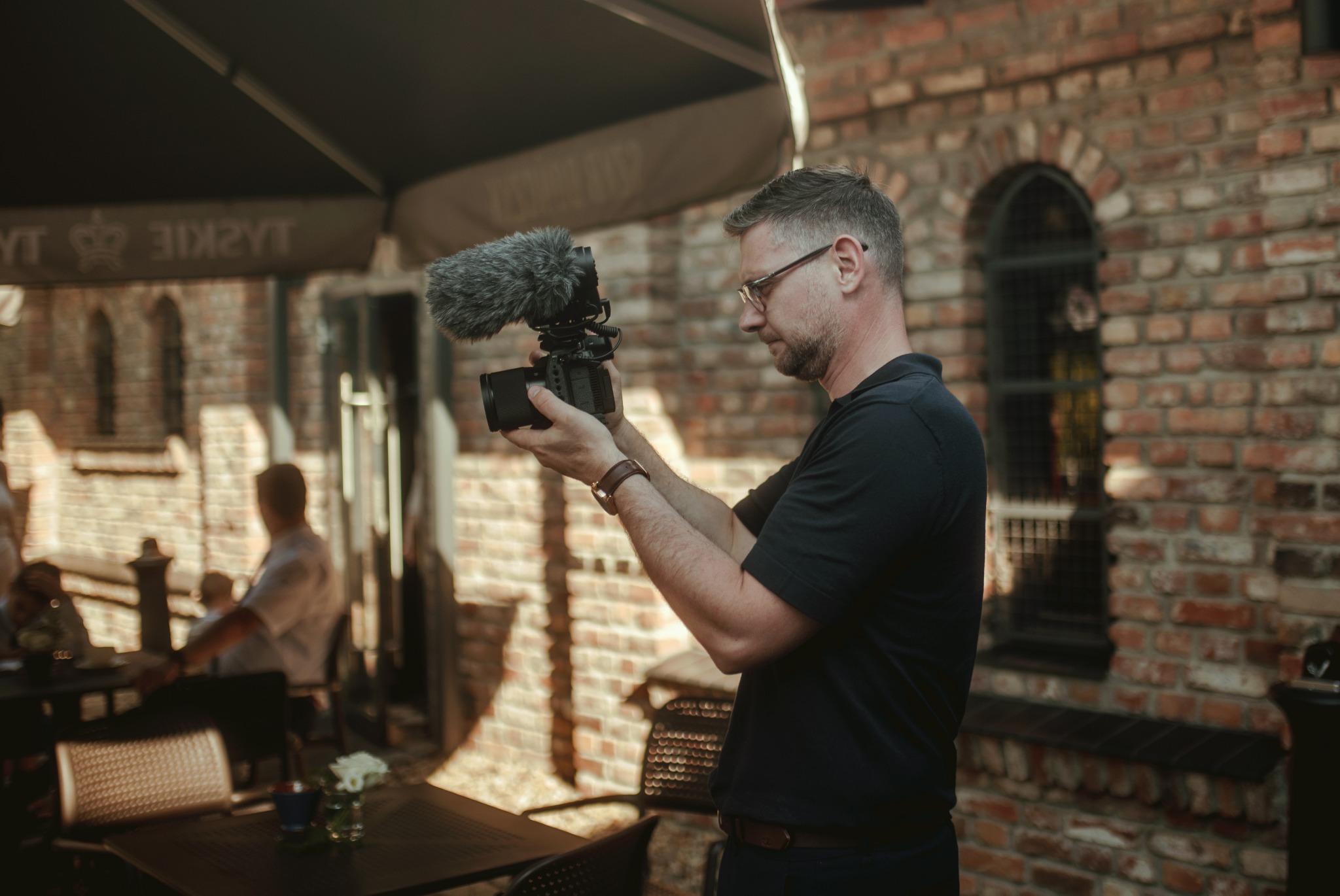 Mężczyzna z brodą i okularach trzyma kamerę z mikrofonem typu shotgun na tle ceglanej ściany i stolików kawiarnianych.