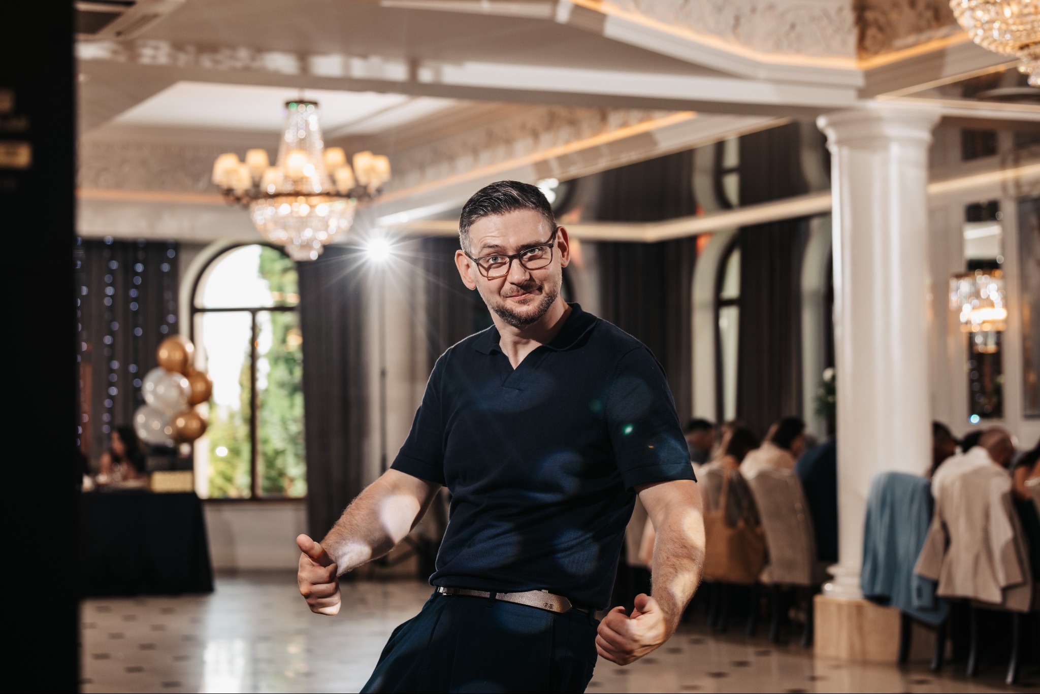 Uśmiechnięty mężczyzna w okularach, w eleganckim wnętrzu z żyrandolem, daje dwa kciuki w górę. W tle goście siedzą przy stołach, widoczne kolumny i dekoracje.